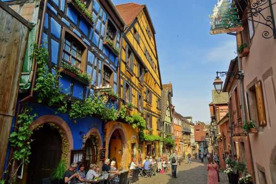 Bunte Fachwerkhäuser in der Altstadt von Riquewihr mit Straßencafé und blumengeschmückten Fassaden – ein typisches Bild des elsässischen Weinortes.