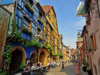 Bunte Fachwerkhäuser in der Altstadt von Riquewihr mit Straßencafé und blumengeschmückten Fassaden – ein typisches Bild des elsässischen Weinortes.